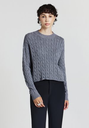 Maglione grigio a trecce con scollatura rotonda, lunghezza cropped e polsini a costine. Indossato con pantaloni neri.