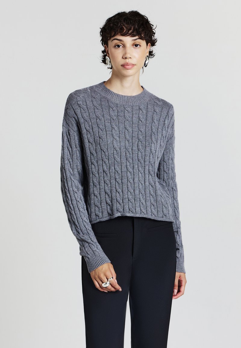 Maglione grigio a trecce con scollatura rotonda, lunghezza cropped e polsini a costine. Indossato con pantaloni neri.
