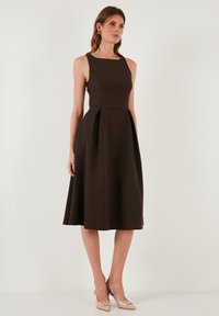 Brązowa sukienka bez rękawów z dopasowanym topem i rozkloszowanym dołem. Gładki, teksturowany materiał; długość do połowy łydki, boczne fałdy i okrągły dekolt.