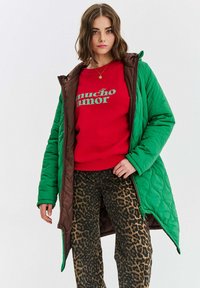 Zelený prešívaný dlhý kabát s hnedou podšívkou, skombinovaný s červeným mikinovým tričkom so zeleným textom a nohavicami s leopardím potlačou so širokými nohavicami.