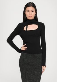 Črna rebrasta turtleneck pulover s izrezom in zapirsko kovinico na prsih, v kombinaciji s sijočo črno svilenko.