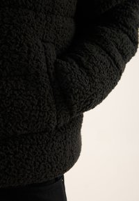 Schwarze Fleecejacke mit strukturierter, flauschiger Oberfläche; verfügt über eine Vordertasche und enganliegende Bündchen für Wärme und Komfort.