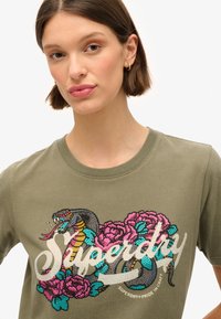 Olijfgroen katoenen T-shirt met een kleurrijke slang en bloemgrafiek met glinsterende accenten. Ronde halslijn en korte mouwen.