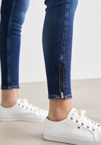 Jeans ajustados de mezclilla en azul oscuro con detalle de cremallera en el tobillo. Los jeans presentan una cintura regular y costuras en contraste. Combinados con zapatillas blancas.