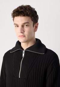 Suéter negro de punto con textura acanalada, que presenta un gran cuello y cremallera de media longitud, diseñado con un patrón geométrico en el frente.