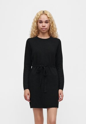 Femme aux longs cheveux blonds bouclés, portant une robe noire à manches longues avec une ceinture nouée à la taille, se tenant devant un fond uni clair.