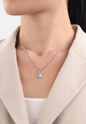 Zilveren ketting met een gestructureerde, klavervormige hanger. De hanger hangt aan een slanke, metalen ketting die mooi aansluit bij een lichtbeige blouse.