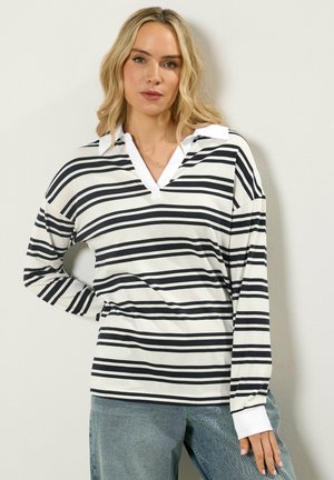 Kobieta o blond włosach, ubrana w biało-czarną pasiastą bluzkę polo na długi rękaw i niebieskie dżinsy, stoi na tle gładkiej ściany.