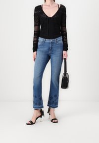 Czarna, koronkowa bluzka z długim rękawem i dekoltem w serek, zestawiona z niebieskimi jeansami o średnim stanie ozdobionymi haftowanymi kwiatami oraz czarnymi sandałami na obcasie.