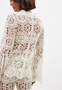 Crochet top featuring a floral pattern in cream with long, flared sleeves and a high neckline. The back shows intricate openwork detailing.