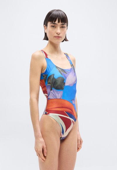 Γυναίκα με κοντά σκούρα μαλλιά που φοράει ένα ολόσωμο μαγιό με πολύχρωμο αφηρημένο σχέδιο, μπροστά σε απλό φωτεινό φόντο.