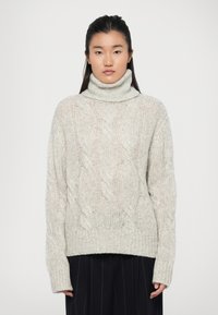 Pull à col roulé en tricot câble gris clair avec ourlet et poignets côtelés. Fait de fil épais et texturé. Le modèle porte en dessous un pantalon plissé foncé.