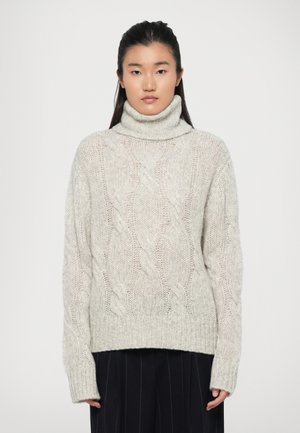 Lys grå kabelstrikket genser med høy hals og ribbestrikket kant og mansjetter. Laget av tykk, strukturert garn. Modellen har på seg mørke, plisserte bukser under.