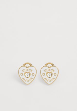 Boucles d'oreilles en forme de cœur dorées et blanches avec le texte "GUESS AMORE" et un cœur en cristal central entouré de deux petits cristaux chacune.