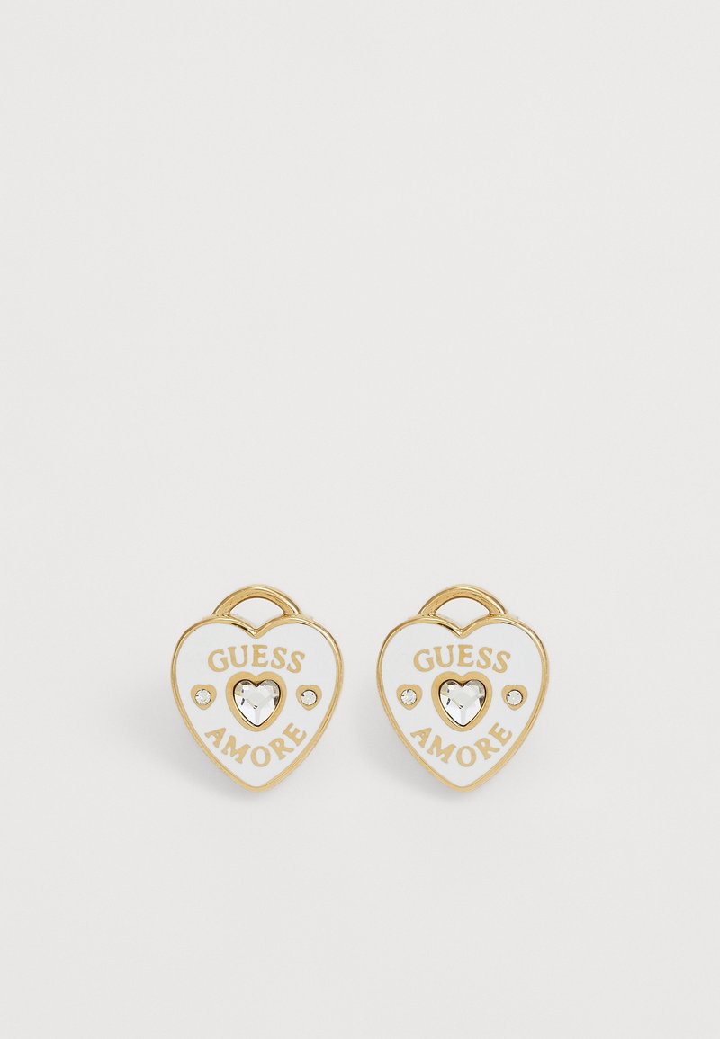 Boucles d'oreilles en forme de cœur dorées et blanches avec le texte "GUESS AMORE" et un cœur en cristal central entouré de deux petits cristaux chacune.