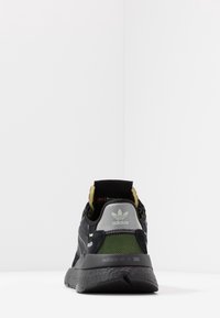 Zapatilla negra de Adidas con parte superior de malla y ante, lengüeta trasera gris, acentos reflectantes y detalle de marca verde. Suela de goma gruesa.