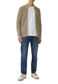 Beige gestrickte Strickjacke mit Stehkragen, über einem weißen T-Shirt getragen, kombiniert mit blauen Jeans und beigen Sneakers.