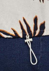 Tessuto bianco con motivo floreale arancione e navy, caratterizzato da un cordoncino con un toggle bianco all'orlo, abbinato a jeans scuri.