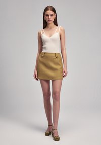 Top bianco a costine con spalline sottili, abbinato a una mini gonna dorata con texture e tasche frontali, accompagnato da scarpe con tacco verde chiaro.