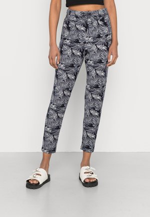 Persoon die zwart-witte broek met tropisch bladerenpatroon en witte sandalen met gesp draagt, staand tegen een effen lichte achtergrond.