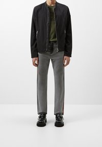 Zwarte bomberjack met ritsen, olijfgroen T-shirt eronder, grijze spijkerbroek met oranje zijstrepen en zwarte gespschoenen.