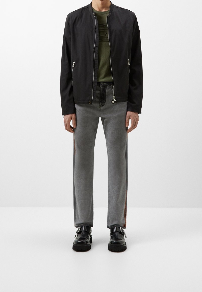 Zwarte bomberjack met ritsen, olijfgroen T-shirt eronder, grijze spijkerbroek met oranje zijstrepen en zwarte gespschoenen.