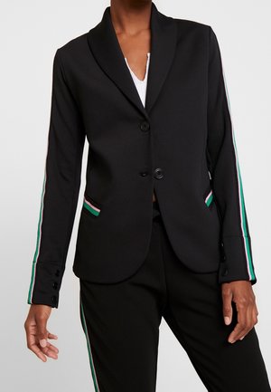 Zwarte blazer met groene, roze en witte strepen op de mouwen en zakranden, gedragen over een wit topje, gecombineerd met bijpassende broek.