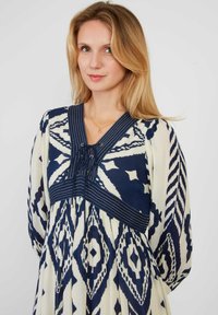 Marineblauwe en witte jurk met een patroon, featuring een V-hals met vetersluiting, een geplooide rok en lange mouwen met contrasterende diagonale strepen.
