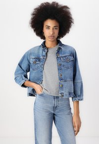 Denim jacket with a faded blue hue, button front, two chest pockets, and rolled sleeves, paired with a grey T-shirt and matching jeans.