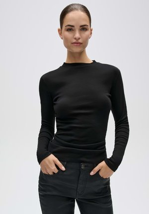 Frau mit dunklen zurückgebundenen Haaren, die ein schwarzes Langarmshirt und schwarze Hose trägt, steht mit den Händen in den Taschen vor einem einfarbigen Hintergrund.