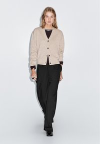Beige stickad cardigan med V-ringning, svarta knappar, lagd över en mörk topp, i kombination med lösa svarta byxor och svarta skor.