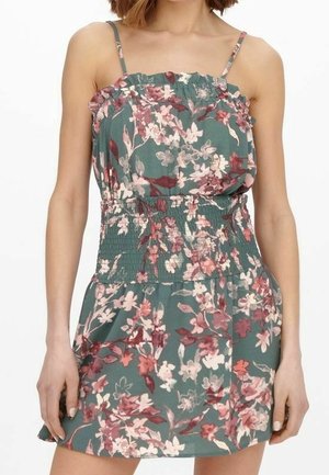 Frau trägt ärmelloses, grünes Kleid mit Blumenmuster, dünnen Trägern, gesmokter Taille und Rüschendetail am Oberteil und Rock.