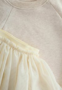 Ljust beige sweatshirt med ribbat halsringning bredvid en krämfärgad tullekjol med samlad elastisk midja, som visar upp mjuka texturer och lager.