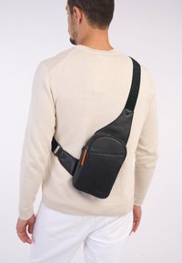 Borsa a tracolla nera in pelle con tracolla regolabile, forma rettangolare, texture liscia e zip arancione, indossata sopra un maglione chiaro.