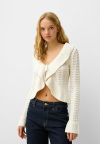 Cardigan en crochet blanc avec un devant ouvert, un grand col et des manches évasées. Associé à un jean bleu foncé taille haute.