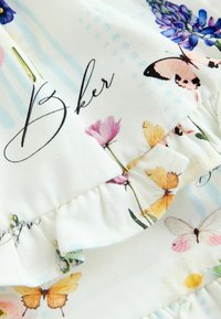 Hvidt stof med farverige sommerfugle- og blomsterprint, sort kursiv tekst med ordet "Bker" og flæset søm.