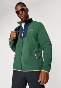 Veste en polaire verte avec un panneau avant bleu marine, poches latérales zippées et finitions beige. Le modèle porte des lunettes de soleil et tient un sac noir.
