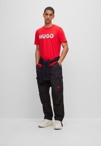 Red t-shirt with "HUGO" logo, paired with black cargo pants featuring pockets and a red accent. White sneakers complete the outfit.