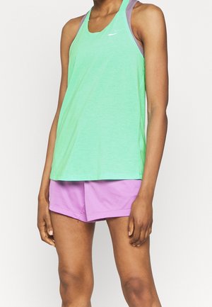 Kvinde iført en mintgrøn ærmeløs træningstop over en lilla sports-bh og lyse lilla shorts, stående mod en ensfarvet baggrund.