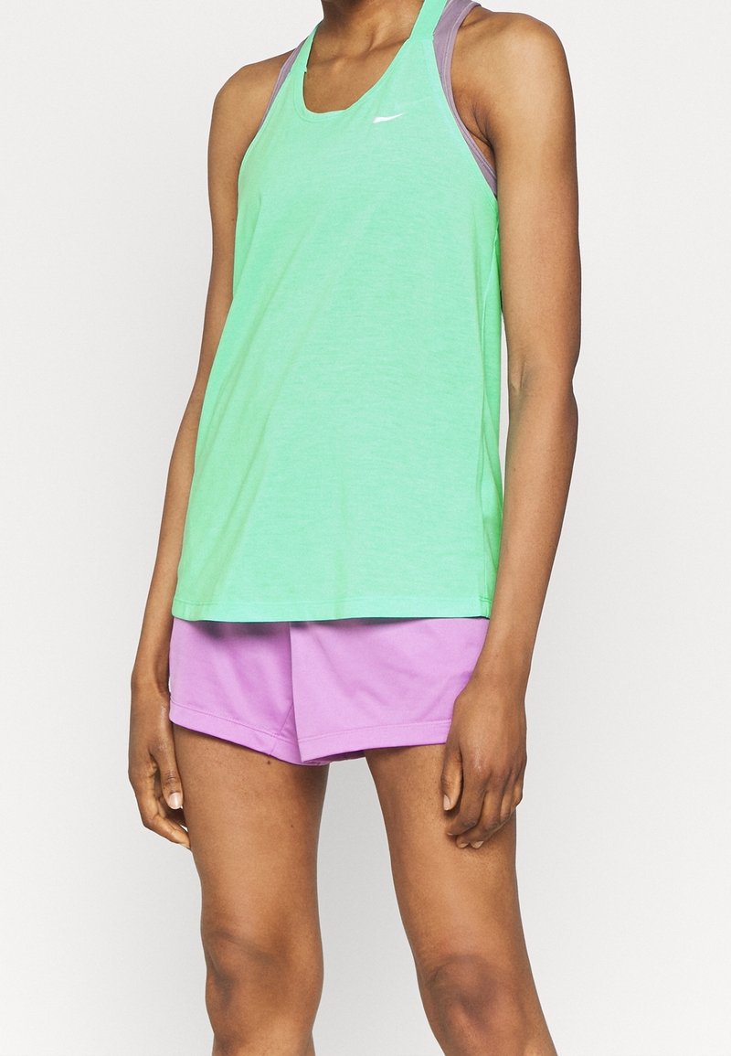 Femme portant un débardeur de sport vert menthe par-dessus un soutien-gorge de sport violet et un short violet clair, debout devant un fond uni.