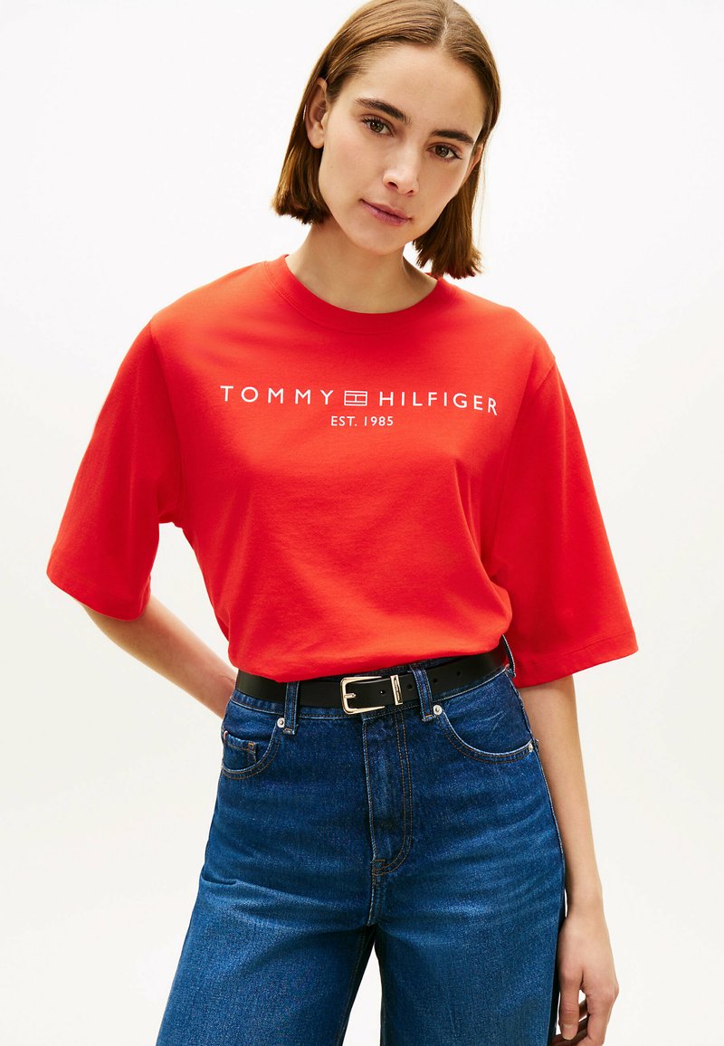 Femme aux cheveux courts bruns portant un T-shirt rouge Tommy Hilfiger et un jean bleu, debout devant un fond blanc.