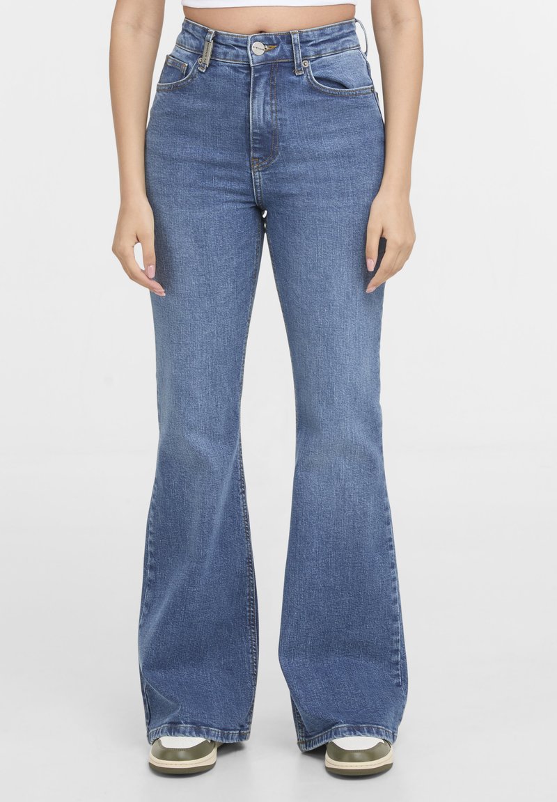 Femme portant un jean évasé bleu taille haute et des baskets blanches, debout devant un fond blanc uni.