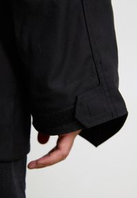 Czarna tkanina kurtki z fakturowanym wykończeniem, z zapięciem na rzep przy mankietach i dopasowanymi szwami w pobliżu nadgarstka.