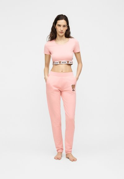Γυναίκα φοράει ροζ crop top Moschino με λωρίδα λογότυπου και ταιριαστό παντελόνι joggers με λογότυπο αρκούδας, στέκεται ξυπόλητη μπροστά σε λευκό φόντο.