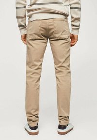 Hombre con pantalones beige, zapatillas blancas con tacones marrones y un suéter beige con rayas horizontales, de pie de espaldas.