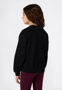 Giovane con lunghi capelli castani, indossa una felpa nera e pantaloni bordeaux, rivolto verso sinistra su uno sfondo bianco uniforme.