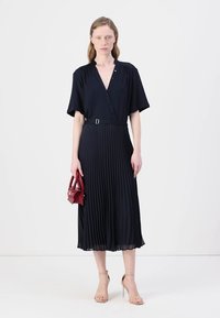 Navy pleated dress with short sleeves, v-neck, and a belt at the waist. Model holding a small red handbag, wearing nude heeled sandals.