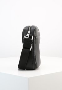 Black leather crossbody bag with a rectangular shape, zip closure, and adjustable textile strap. Smooth texture and minimalistic design.
