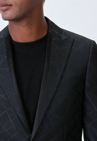 Schwarze Blazer mit einem subtilen geometrischen Muster, ausgestattet mit einem Satin-Schalkragen und einer strukturierten Passform, getragen über einem schwarzen Rundhalshemd.