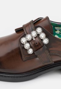 Chaussure en cuir marron avec une lanière décorative ornée de huit grandes pierres précieuses claires et une couture détaillée sur une semelle noire.